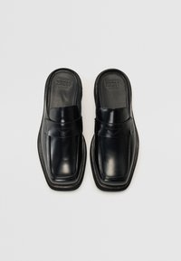 Zapatos negros de cuero deslizables con punta cuadrada, suela plana y una correa frontal con detalles de costura para un diseño adicional.