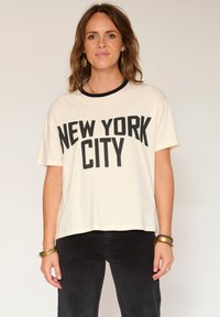 Femme portant un T-shirt blanc avec le texte noir "NEW YORK CITY", un pantalon noir, des boucles d'oreilles en or et des bracelets, se tenant devant un fond clair.