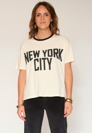 Femme portant un T-shirt blanc avec le texte noir "NEW YORK CITY", un pantalon noir, des boucles d'oreilles en or et des bracelets, se tenant devant un fond clair.