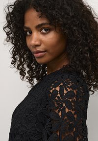 Black lace top with intricate floral embroidery, sheer sleeves, and a textured fabric. Close-up view focuses on details and patterns.