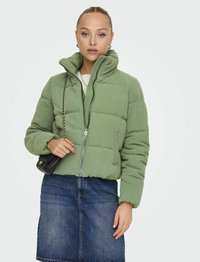 Veste matelassée en velours côtelé vert avec un col haut, fermeture éclair et poignets côtelés. Associée à une jupe en denim. Tenant un sac à main noir.