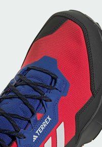 Punaiset ja mustat urheilukengät, joissa on teksturoitu päällinen, siniset yksityiskohdat, nauhakiinnitys ja vahvistettu varvaskorkki. Brändin nimi näkyy.