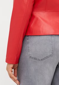 Κόκκινο τεχνητό δερμάτινο μπουφάν με εφαρμοστή σχεδίαση. Το μανίκι έχει μια δομημένη μανσέτα; φορεμένο πάνω από ανοιχτό γκρι τζιν με μία μόνο πίσω τσέπη.