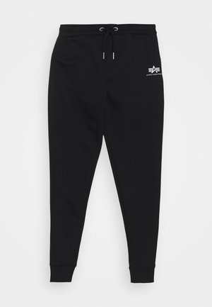 Pantalones deportivos negros ajustados con cintura de cordón y logo blanco de Alpha Industries en la cadera izquierda, puños acanalados en los tobillos, disposición en plano sobre fondo blanco.