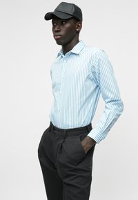 Cămașă albastră deschis cu dungi albe, cu guler tip button-down, asortată cu pantaloni negri tapered. O șapcă neagră adaugă un detaliu contrastant.
