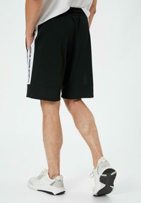 Sorte træningsshorts med elastisk talje, der har sidelommer og hvide tekstdetaljer på de ydre sømme. Bæres med lyse sneakers.