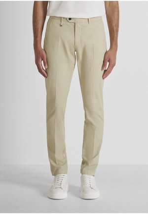 Uomo che indossa pantaloni beige slim fit e sneakers bianche, in piedi contro uno sfondo chiaro e uniforme.