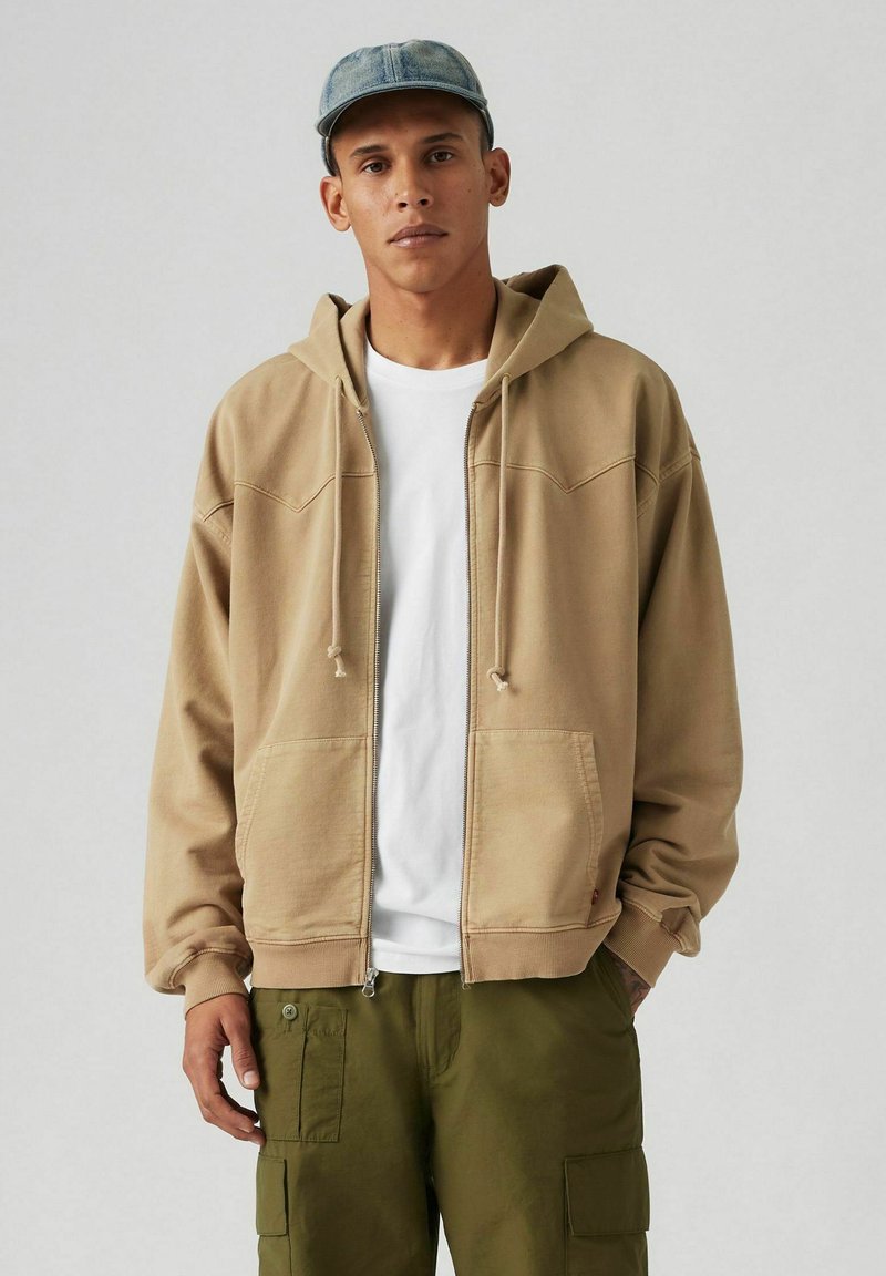 Kaptrui van beige katoen met een rits aan de voorkant, twee voorkanten zakken en een trekkoord. Witte t-shirt eronder, gecombineerd met olijfgroene cargobroek.