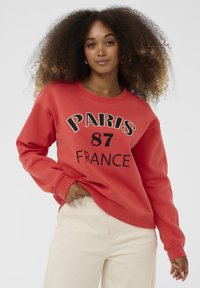 Ženska s kodrastimi lasmi, oblečena v rdečo majico s napisom "PARIS 87 FRANCE" in kremo barvne hlače, ki pozira pred enobarvno svetlo ozadjem.