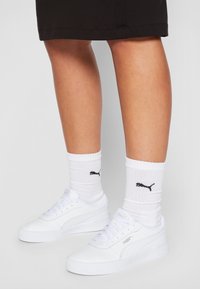 Sneakers bianchi con suola in gomma testurizzata, dotati di tomaia sintetica liscia e chiusura con lacci, abbinati a calzini bianchi a costine con un logo nero.