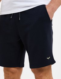 Kraťasy do kolen v námořnické modři vyrobené z měkké bavlny, s elastickým pasem na šňůrku a malým zeleným logem na dolní části nohy.