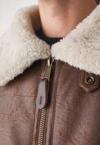 Hnědá kožená bunda s bílým, chlupatým umělým kožešinovým límcem. Má zlatý zip a hnědou koženou táhlo, spolu s detailním knoflíkem.