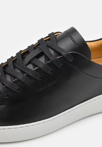 Sneaker de couro preto com atacadores pretos trançados, superfície lisa, sola de borracha branca e marcação perto do calcanhar. Interior amarelo mostarda.