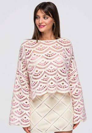 Mujer con cabello castaño liso lleva un top de encaje con borde en forma de conchas en blanco y rosa con mangas anchas y una falda crema con patrón de encaje en forma de diamante.