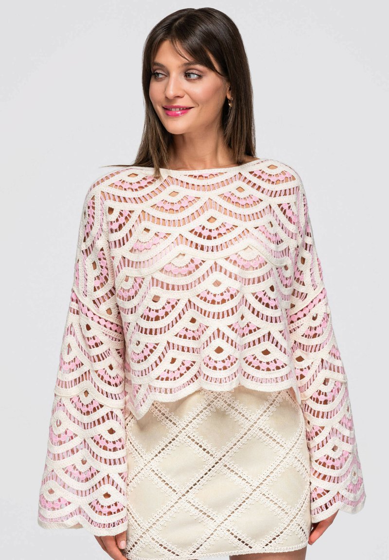 Femme aux cheveux bruns et raides portant un haut en dentelle blanche et rose à bord festonné avec de larges manches et une jupe crème avec un motif en dentelle losange.