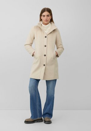Manteau en laine beige avec capuche, présentant un devant à boutons, des poches latérales et des poignets côtelés. Porté sur un col roulé blanc et associé à un jean évasé.