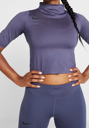 Kobieta ubrana w fioletowy crop top Nike i dopasowane legginsy z wysokim stanem stoi z rękami na biodrach, pokazując szczegóły odzieży sportowej.