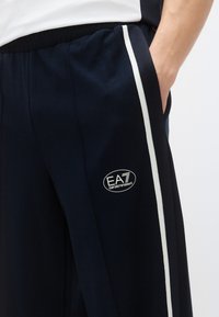 Tmavomodré športové nohavice s bielymi bočnými pruhmi, s oválnou nášivkou s logom "EA7 Emporio Armani" na ľavom stehne.