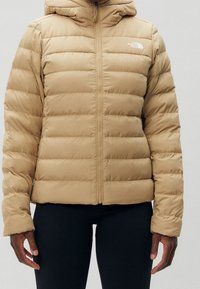 Doudoune beige avec capuche, présentant des coutures horizontales, une fermeture éclair à l'avant et un détail de logo sur le côté gauche de la poitrine. Tissu lisse et isolant.