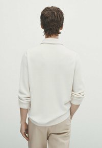 Pull côtelé beige clair avec col à revers et manches retroussées, vu de dos, assorti à un pantalon beige. Détails du tissu texturé visibles.