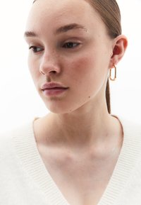 Orecchino a cerchio in oro con forma rettangolare, caratterizzato da una superficie liscia e lucida. Il modello indossa un maglione a V di colore chiaro.