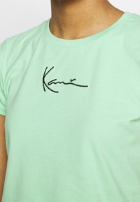 Persona que lleva una camiseta verde menta de manga corta con texto en cursiva negra en el pecho.