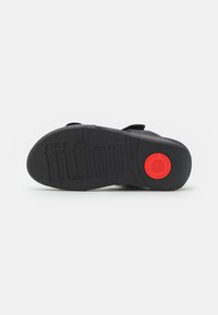 Černá guma na podrážce sandálů s texturovaným gripem, obsahující červený kruhový akcent a vyražené logo "fitflop". Hladký povrch, robustní design.