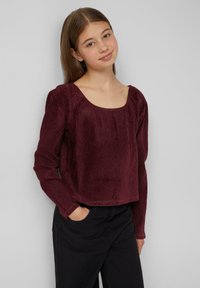 Bordeaux langarmtop met een geribbelde textuur en een brede ronde halslijn, gecombineerd met zwarte broek. Het topje heeft een cropped ontwerp.