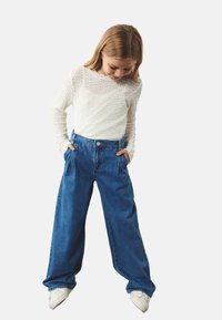 Witte gestructureerde langemouwen top gecombineerd met high-waisted, wijde blauwe denim broek. Model staat met handen in de zakken, draagt witte sneakers.