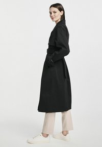 Czarny trenchcoat z paskiem w talii, dużym kołnierzem i guzikami. Model nosi go z beżowymi spodniami i białymi sneakersami.