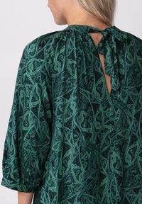 Blusa verde con patrón y detalle de espalda con ojal, escote fruncido y mangas 3/4, que presenta un diseño de cachemira en diferentes tonos de verde.