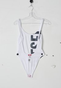 Maillot de bain une pièce blanc avec un dos plongeant, arborant un grand logo noir à l'avant, des fermetures à pression sur les côtés et une étiquette de marque fixée.