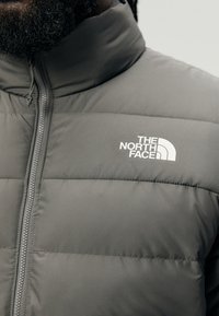 Grå isolert jakke med høy krage, vertikal quiltet design, og en hvit logo på venstre brystområde. Glidelås foran.