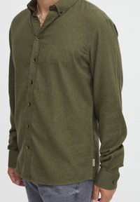 Cămașă cu mânecă lungă, de culoare verde măslinie, fabricată dintr-un material texturat, având un guler butonat și nasturi din lemn pe toată lungimea plăcii frontale.