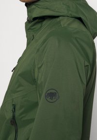 Zielona wodoodporna kurtka z matowym wykończeniem, kieszenią na zamek i bocznym panelem. Na lewym rękawie znajduje się okrągłe logo mamuta.