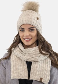 Bonnet tricoté beige clair avec un pompon en fausse fourrure et une écharpe assortie présentant un motif texturé. Matériau doux semblable à de la laine.