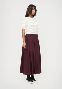 Haut à rayures blanches et beiges à manches courtes avec un col rond associé à une jupe maxi plissée bordeaux. Le mannequin porte des collants noirs et des chaussures à talons pointus.