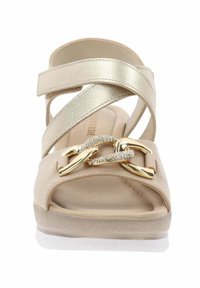 Sandali beige con cinturini incrociati dorati metallici, suola piatta e una fibbia decorativa in oro e strass sulla punta. Texture liscia.