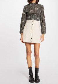 Schwarze Bluse mit floralem Muster und langen Ärmeln, kombiniert mit einem beigen Wildleder-Minirock mit Knopfverschluss und schwarzen Chelsea-Stiefeln.
