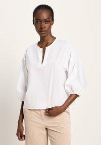 Eine Frau trägt eine weiße, locker sitzende Bluse mit Ballonärmeln und beige Cordhosen und steht vor einem schlichten, hellen Hintergrund.