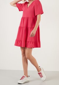 Růžové tiered šaty s výstřihem do V a krátkými nabíranými rukávy, vyrobené z lehké tkaniny, v kombinaci s bílými teniskami a červenými tkaničkami.