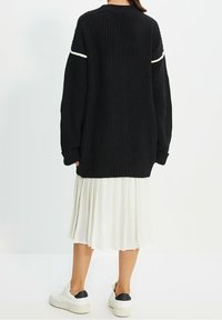 Svart stickad oversize-tröja med vita detaljer på ärmarna, i kombination med en vit plisserad kjol och chunkiga vita sneakers med svart klack.