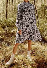 Femme portant une robe ample à manches longues à imprimé léopard et des bottes blanches à la cheville, debout dans une clairière ensoleillée de la forêt.