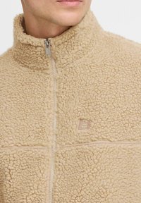 Chaqueta de forro polar beige con un alto cuello y un cierre completo. Presenta una superficie texturizada y un pequeño logo en el lado izquierdo del pecho.