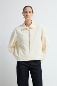 Crème-kleurige ritsjack met een puntige kraag en een relaxed fit. Gemaakt van een gestructureerde stof, gecombineerd met donkere, slim-fit jeans.