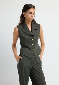 Gilet sans manches de couleur vert foncé, cintré, avec un col cranté, cinq boutons dorés et des poches latérales, en tissu texturé.