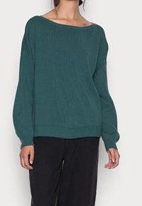 Grüner gerippter Strickpullover mit einem schulterfreien Ausschnitt, lockerer Passform, langen Ärmeln und einem breiten Saum. Getragen mit dunklen Hosen.