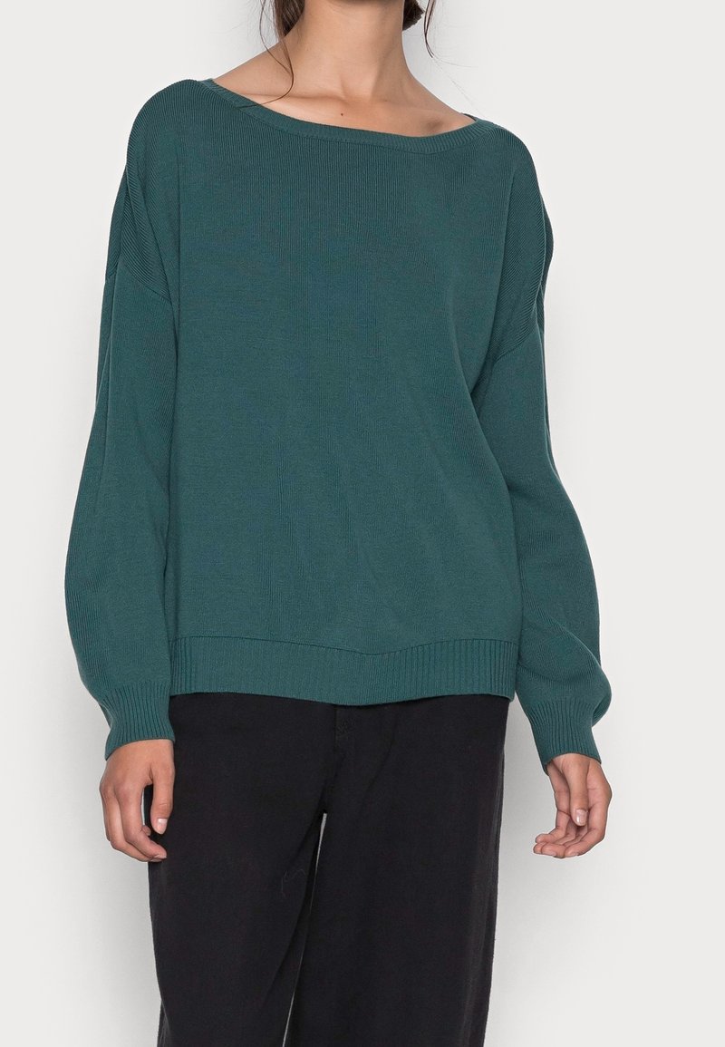 Grüner gerippter Strickpullover mit einem schulterfreien Ausschnitt, lockerer Passform, langen Ärmeln und einem breiten Saum. Getragen mit dunklen Hosen.