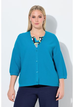 Donna che indossa un cardigan azzurro brillante con bottoni sopra una maglietta a fantasia, abbinato a pantaloni blu scuro, in piedi su uno sfondo bianco.