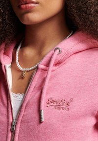 Mujer vistiendo una sudadera rosa de SuperDry sobre una blusa blanca con bordes de encaje, y un collar de perlas con un pequeño colgante de oso de oro.